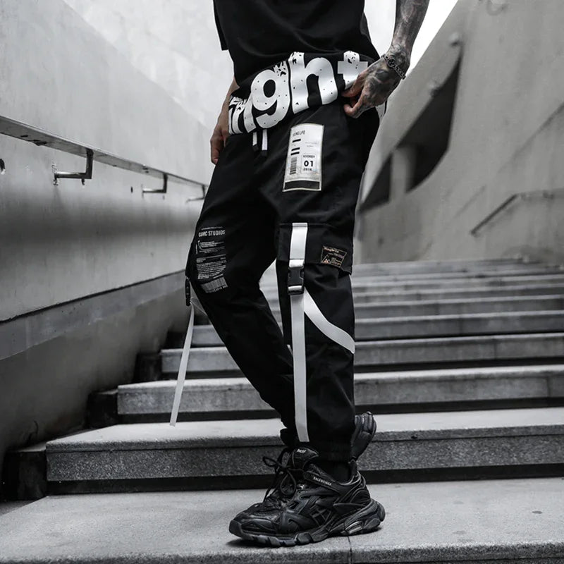 Man wearing black streetwear pants with white straps and patches, standing on concrete stairs