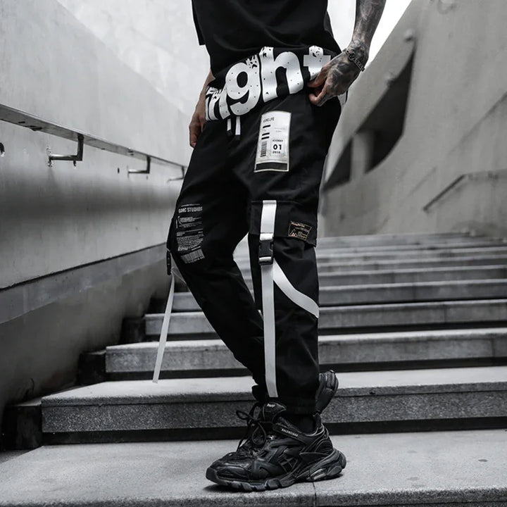 Man wearing black streetwear pants with white straps and patches, standing on concrete stairs