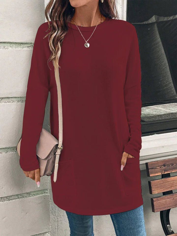 Woman wearing long-sleeve burgundy tunic sweater with pockets and beige crossbody bag by concrete wall