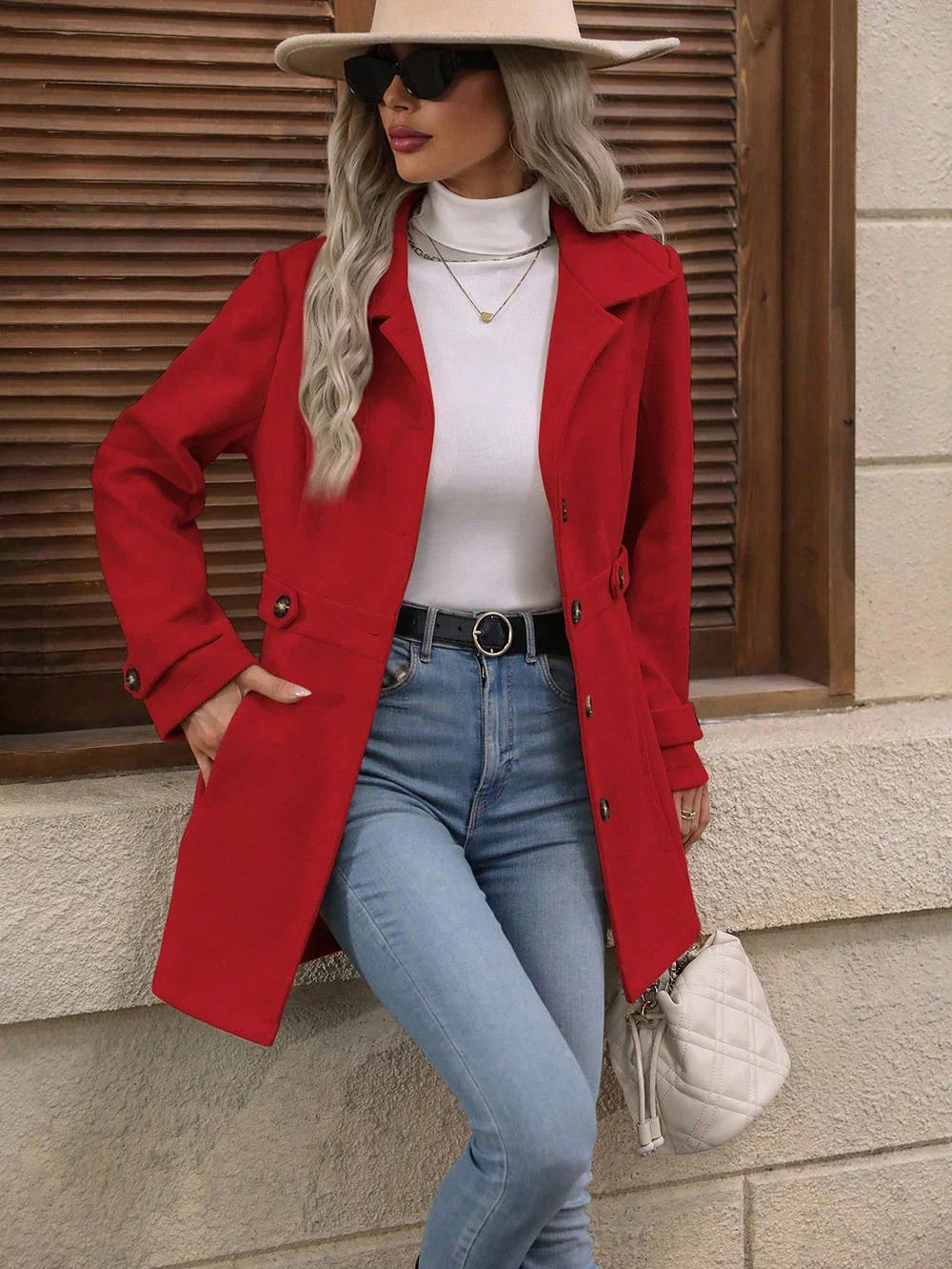 Woman wearing red coat with white turtleneck, light blue jeans, beige hat, and sunglasses, holding white quilted handbag