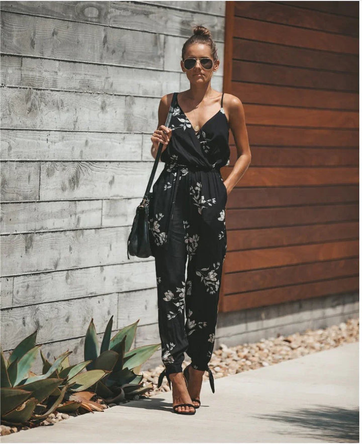 Woman wearing black floral jumpsuit and black heels standing against concrete and wood wall outdoors