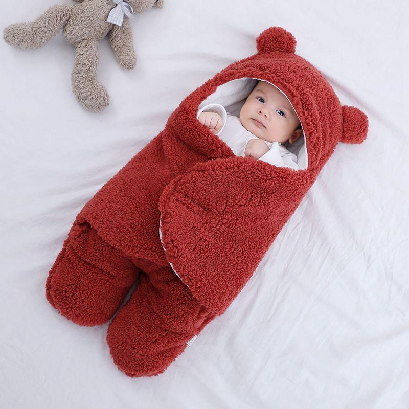 Baby wrapped in a cozy red teddy bear fleece swaddle blanket with ears, lying on white bedding