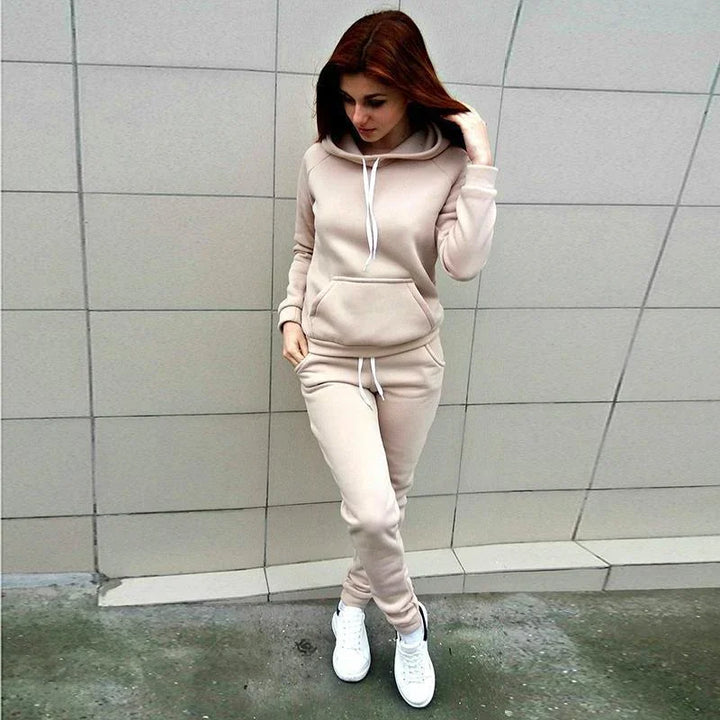 Woman in beige hoodie and matching sweatpants standing against tiled wall in casual outfit with white sneakers