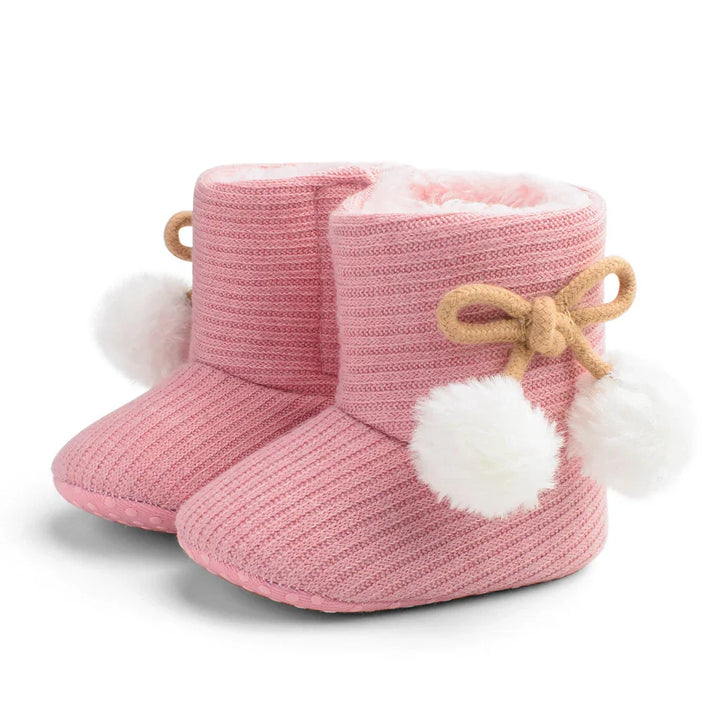 Pair of pink knit baby booties with white fluffy pom-poms and beige bow tie accents