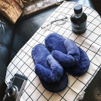 Pair of blue fuzzy slippers on a white grid-patterned cloth with a camera, glasses, and a black bottle on a black leather surface