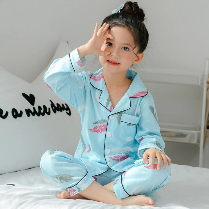 Young girl sitting cross-legged on bed in light blue feather print pajamas, making OK gesture