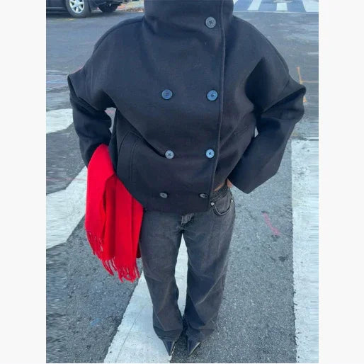 Person wearing oversized black double-breasted coat, dark jeans, pointed black shoes, holding bright red scarf outdoors