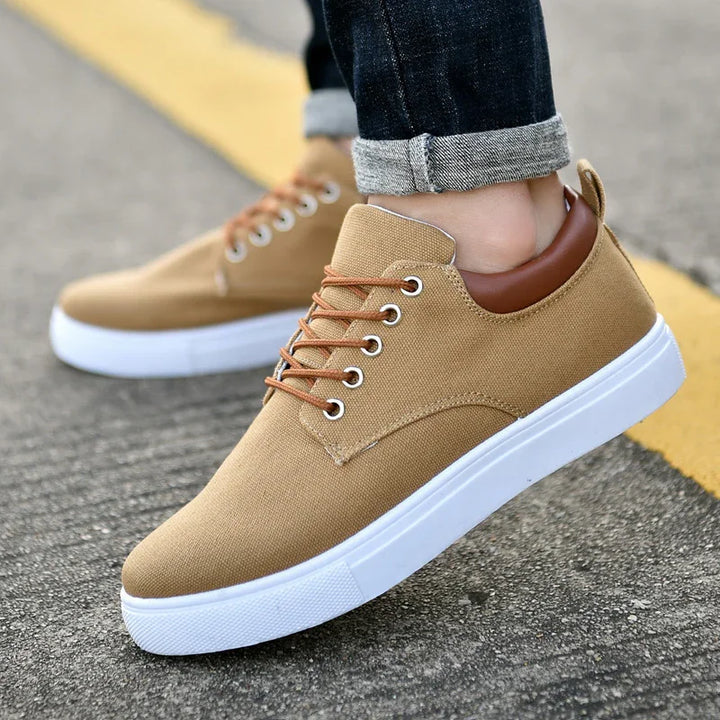 Casual beige canvas sneakers with brown leather accents and white soles worn with rolled-up dark jeans on asphalt