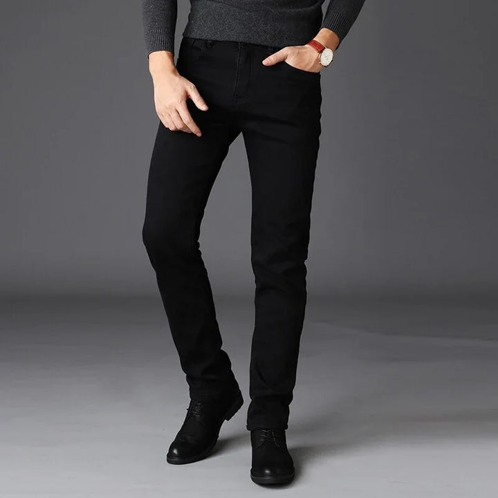 Man wearing slim fit black jeans with black shoes and a dark sweater against gray background