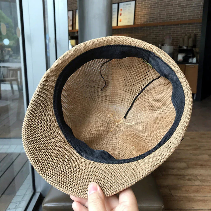 Hand holding a woven straw hat with black inner band in a cafe setting with wooden floor and glass windows