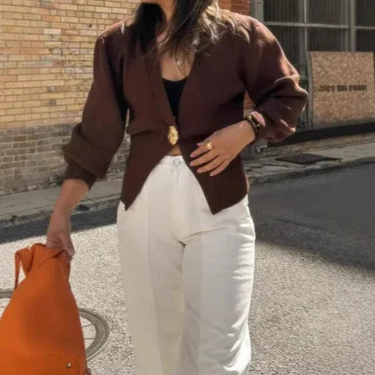Woman wearing brown blazer with gold button and white pants holding orange bag outdoors
