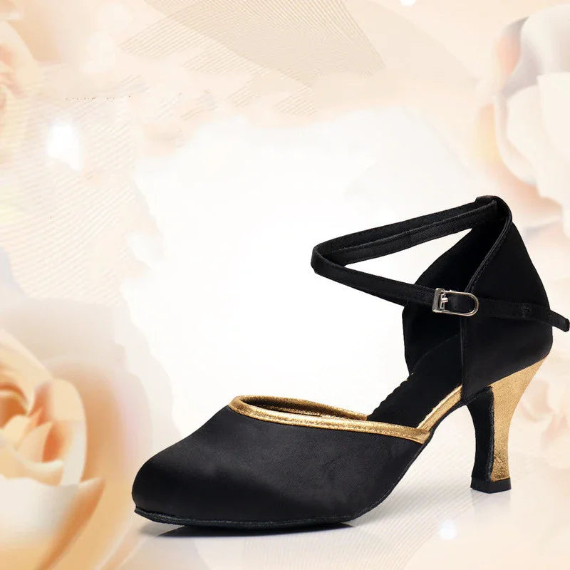 Black satin dance shoe with gold trim, ankle strap, and chunky mid heel on floral background