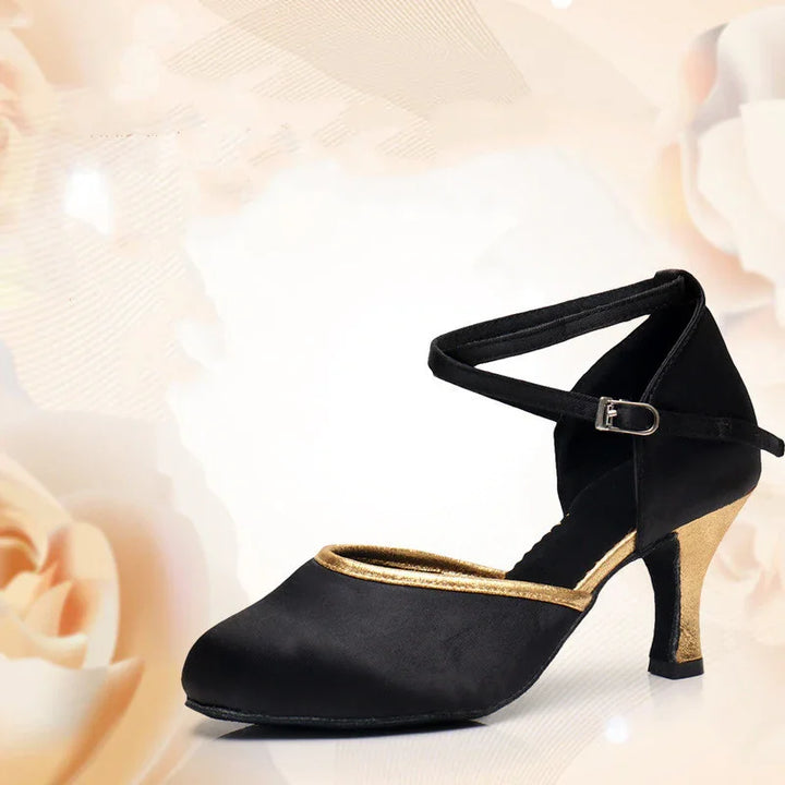 Black satin dance shoe with gold trim, ankle strap, and chunky mid heel on floral background