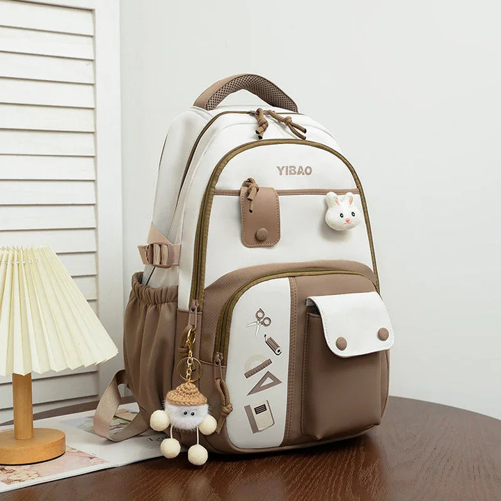 Stylish white and brown YIBAO backpack with school supplies print and cute keychain on wooden table