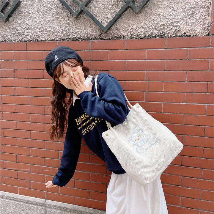 Young woman in navy sweater and beret with white tote bag featuring a cute bear design against a brick wall