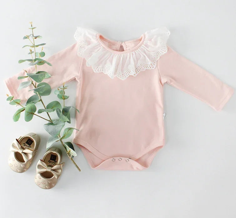 Pink baby onesie with white lace collar, beige baby shoes with bows, and eucalyptus branch on white background