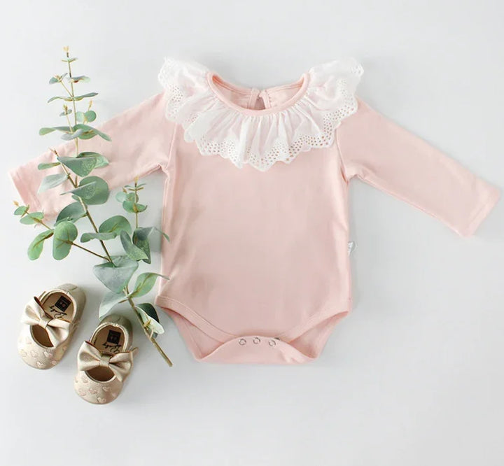 Pink baby onesie with white lace collar, beige baby shoes with bows, and eucalyptus branch on white background