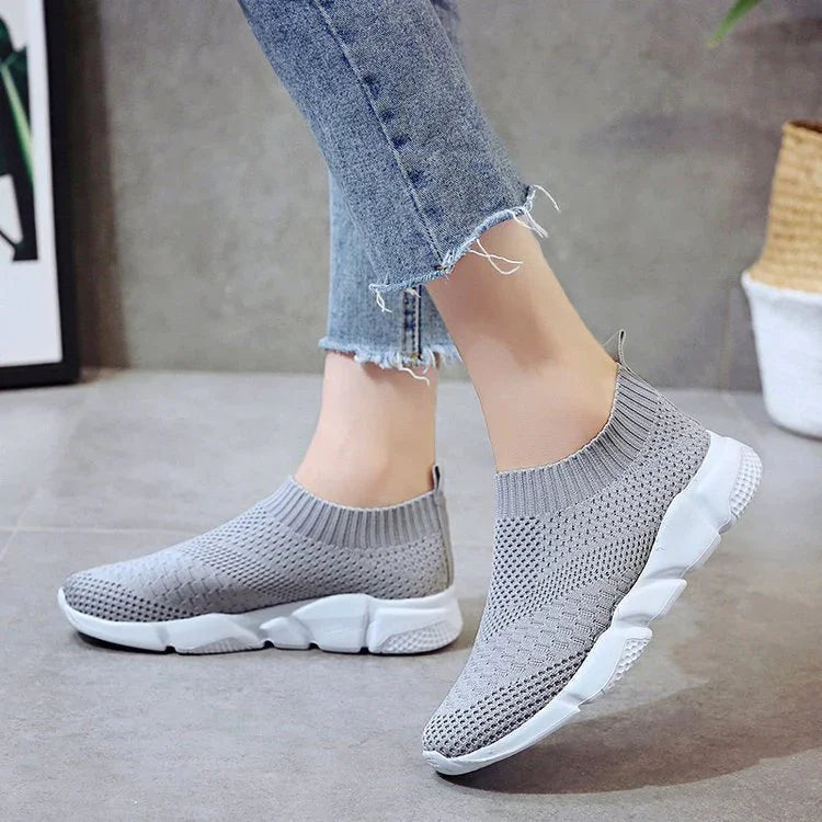 Gray knit slip-on sneakers with white cushioned soles worn with frayed hem denim jeans on gray floor