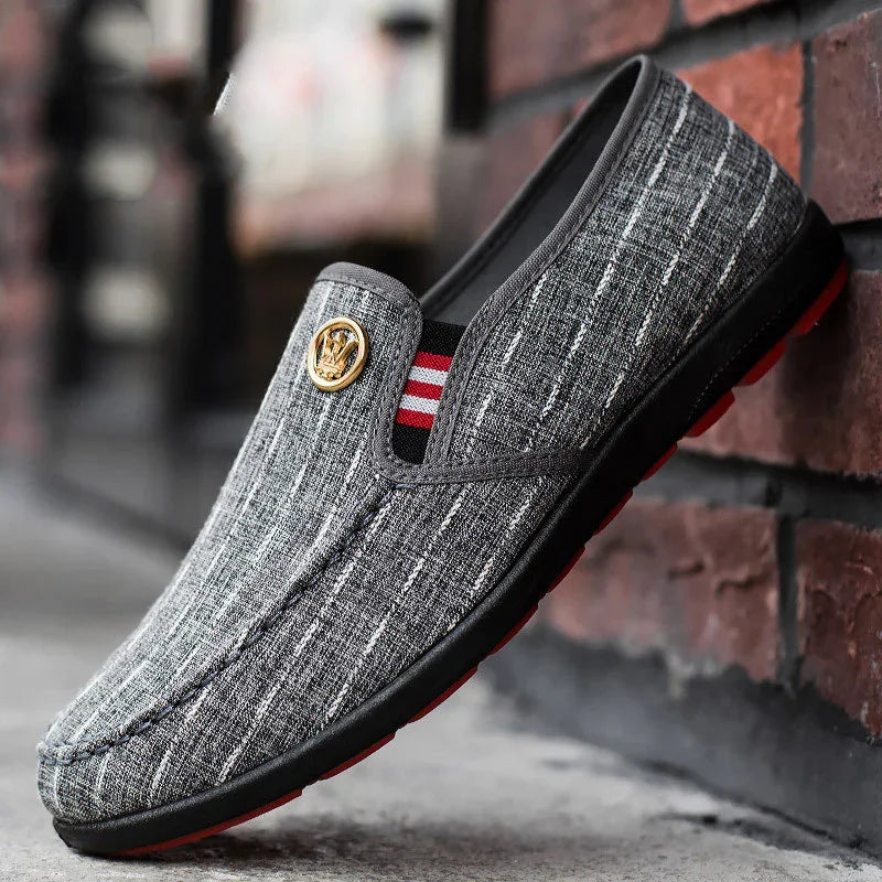 Gray casual men's loafer with textured fabric and gold emblem detail against brick wall