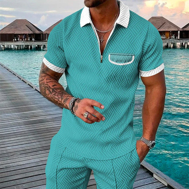 Man wearing turquoise patterned short-sleeve polo shirt and matching pants on wooden pier by tropical overwater bungalows