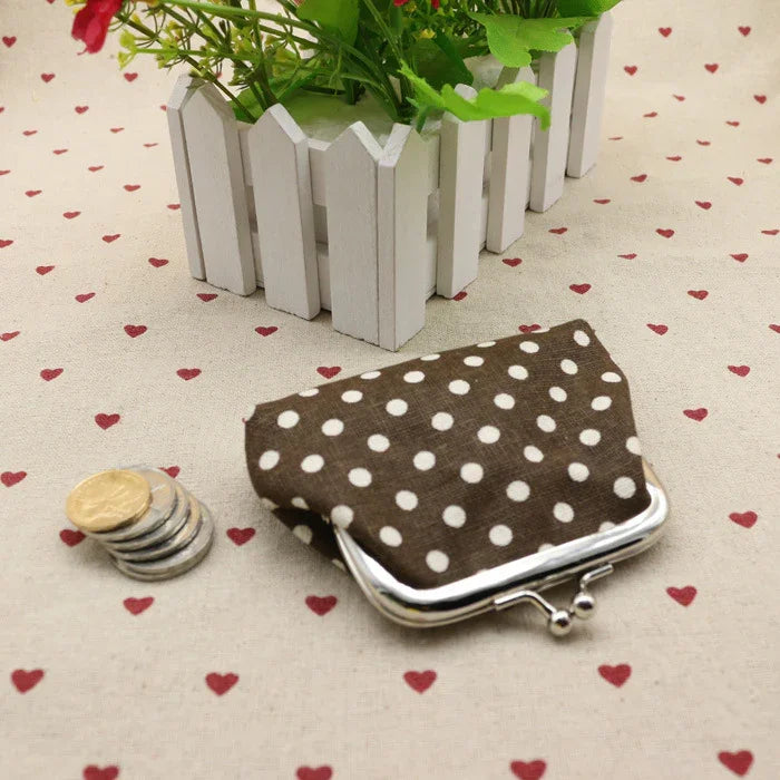 Brown polka dot coin purse with metal clasp on heart-patterned fabric near stacked coins and white picket fence planter with green plants