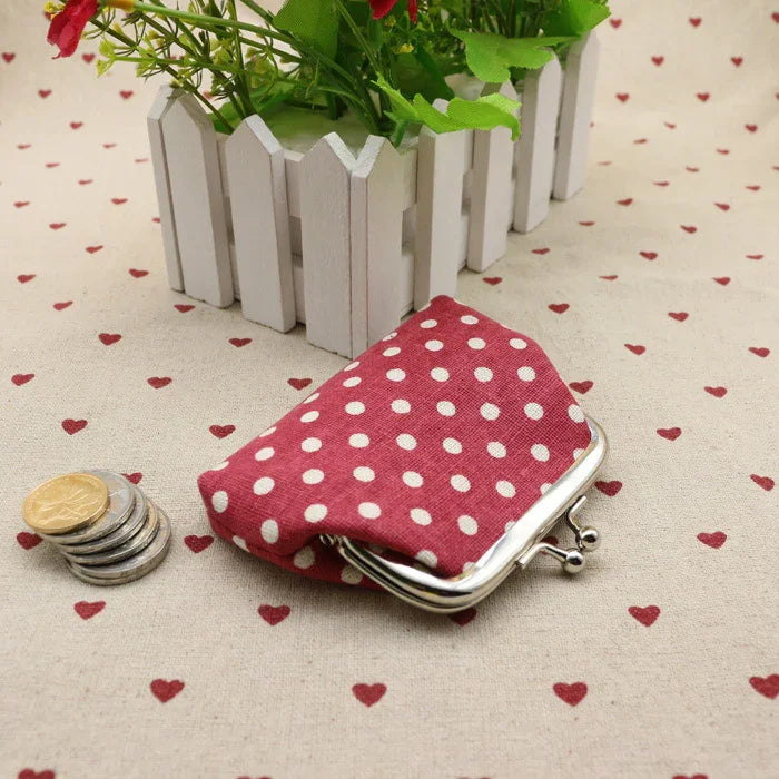 Red polka dot coin purse with metal clasp on heart-patterned tablecloth near stacked coins and white wooden planter with green plants