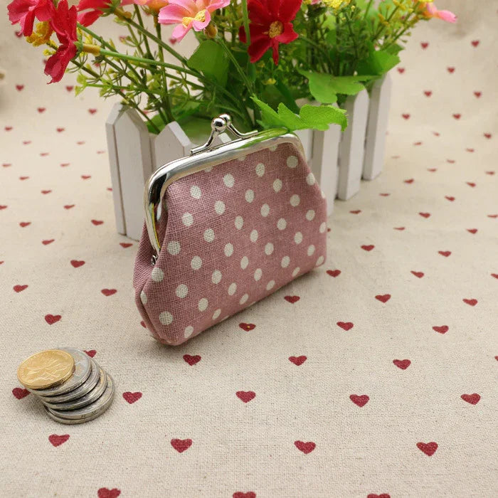 Pink polka dot coin purse with silver clasp on heart-patterned fabric near a small white fence planter with colorful flowers and stacked coins