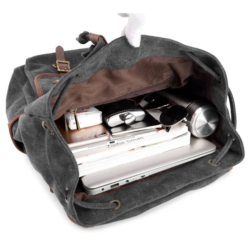 Open dark grey canvas backpack with brown leather accents showing a laptop, notebooks, phone, sunglasses, and stainless water bottle inside