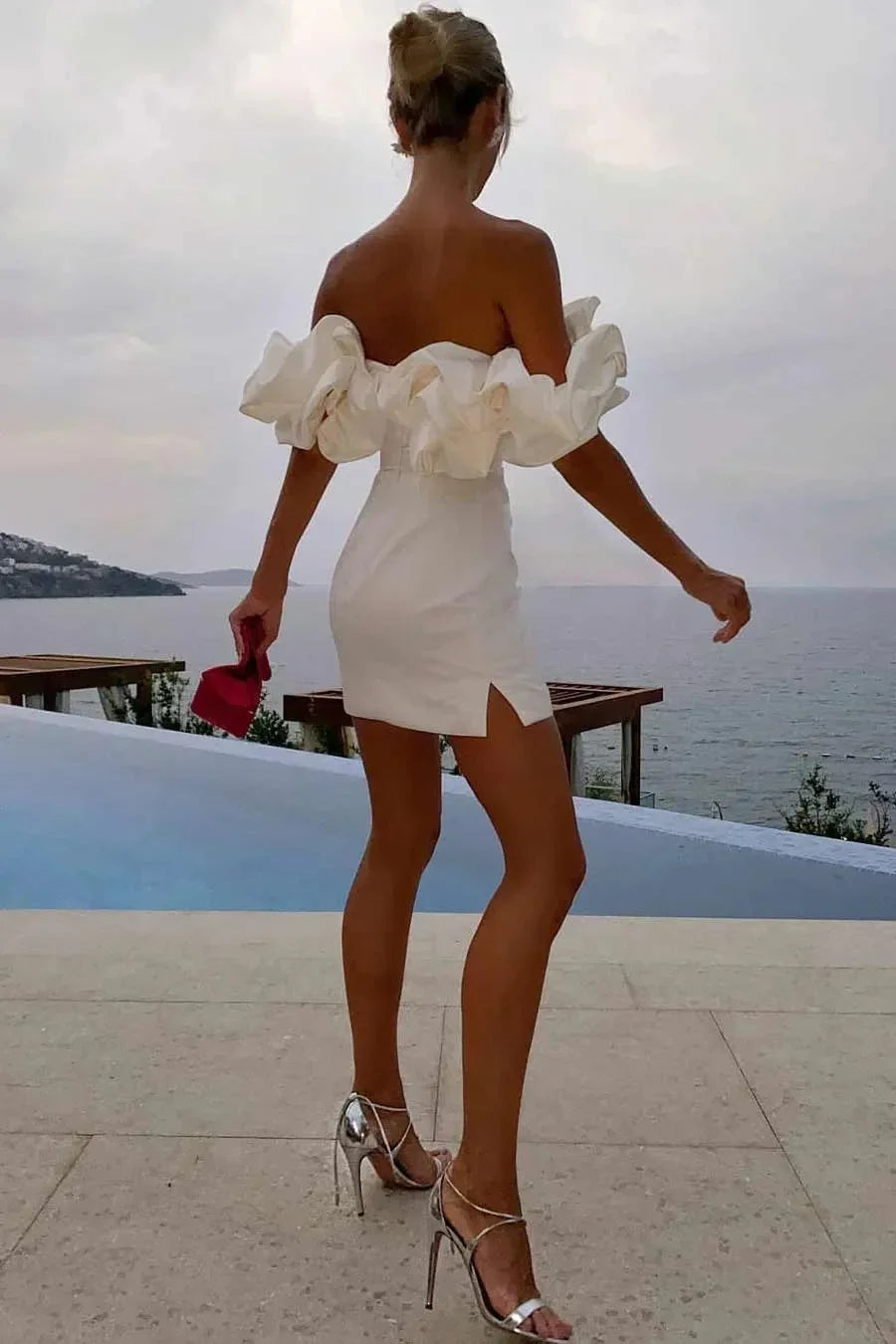 Woman in white off-shoulder dress and silver heels standing by pool overlooking ocean