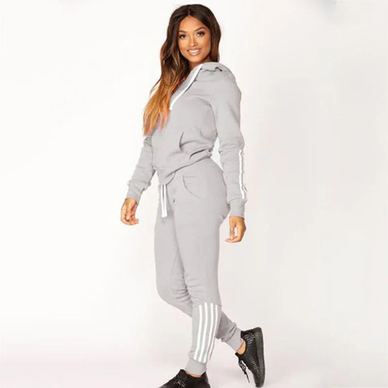 Woman in light gray hooded tracksuit with white stripes, black sneakers, posing against white background