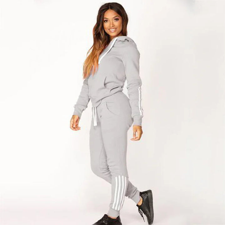 Woman in light gray hooded tracksuit with white stripes, black sneakers, posing against white background