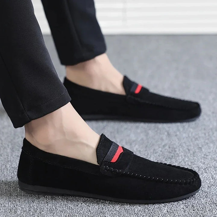 Men's black suede loafers with red detail, worn with black pants on gray carpet