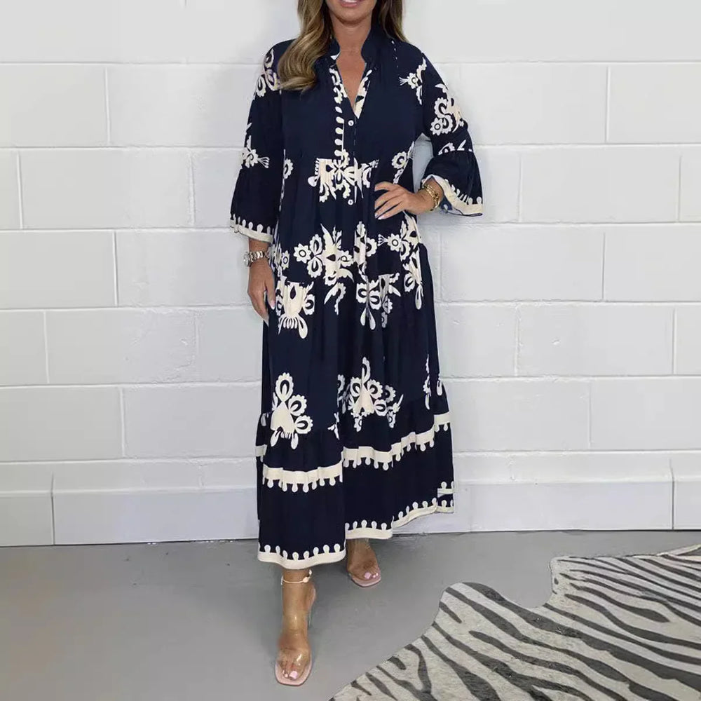 Woman wearing navy blue maxi dress with white floral embroidery and flared sleeves standing by white wall
