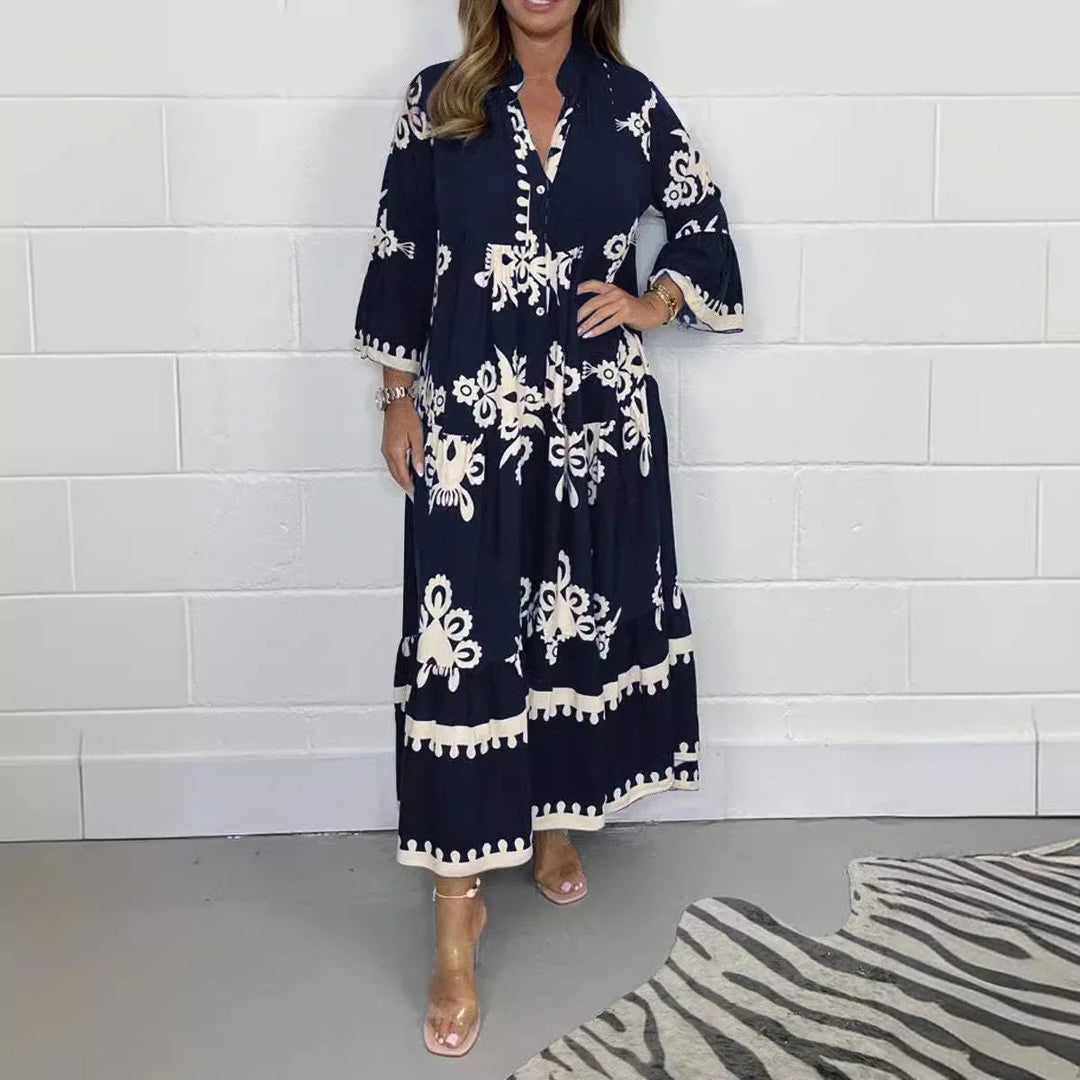 Woman wearing navy blue maxi dress with white floral embroidery and flared sleeves standing by white wall