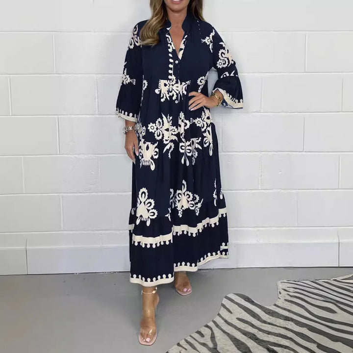 Woman wearing navy blue maxi dress with white floral embroidery and flared sleeves standing by white wall