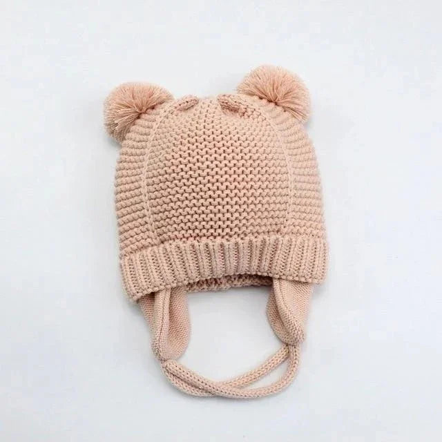 Soft pink knitted baby hat with ear flaps and pom pom ears on white background