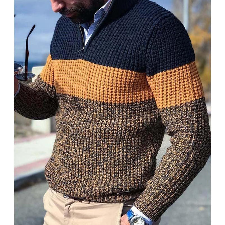 Man wearing color-block knit sweater with navy, mustard, and brown stripes, holding sunglasses outdoors