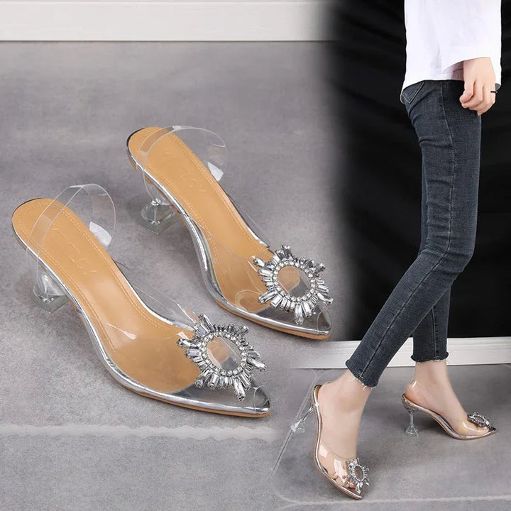Transparent high heel mules with rhinestone decoration worn by person in black jeans