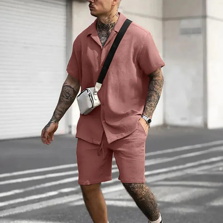 Man with tattoos wearing a matching rose pink short sleeve shirt and shorts set, carrying a white crossbody bag