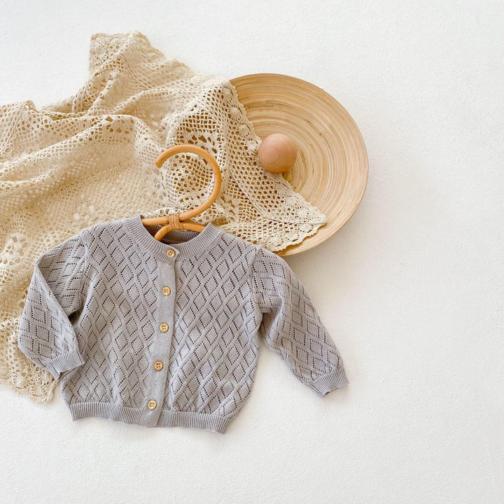 Soft gray baby cardigan with diamond pattern and wooden buttons on a hanger, lace cloth and wooden bowl