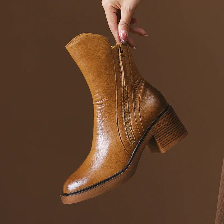 Hand holding a tan leather ankle boot with side zipper and block heel on brown background