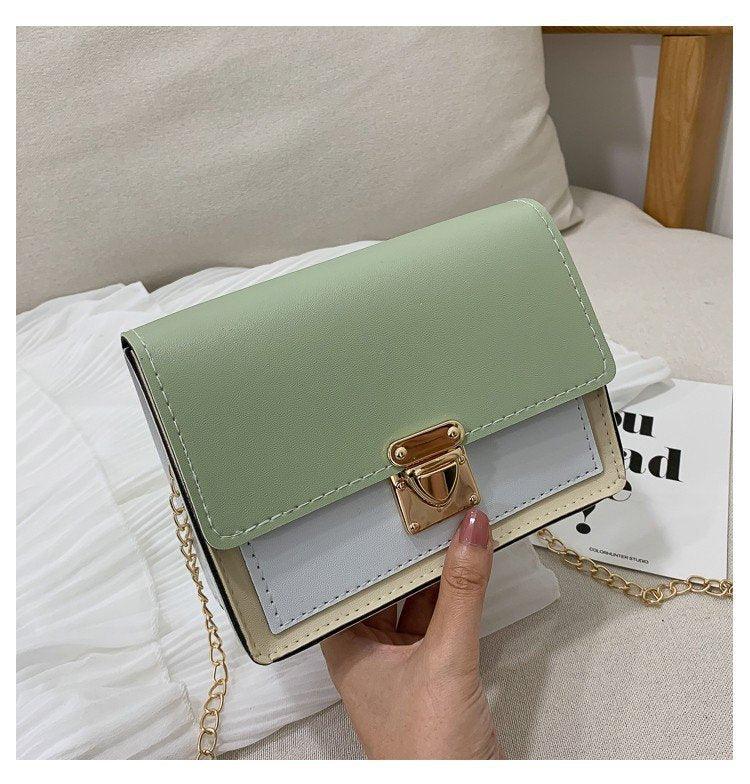Hand holding a stylish small handbag with green flap, white body, and gold metal clasp on beige fabric
