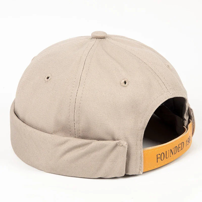 Beige cotton cap with rolled brim and adjustable yellow strap labeled Founded 1985