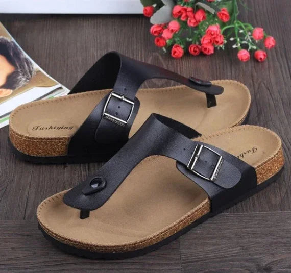 Black leather buckle thong sandals with cork footbed on wooden floor with red flowers