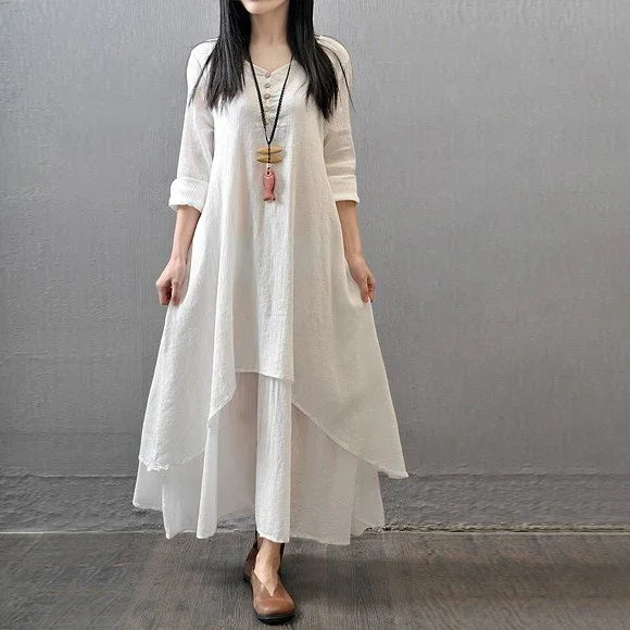 Woman wearing a flowing white layered dress with 3/4 sleeves and brown shoes against gray wall