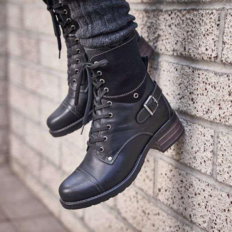 Black leather lace-up ankle boots with buckle detail worn with dark socks against a textured brick wall
