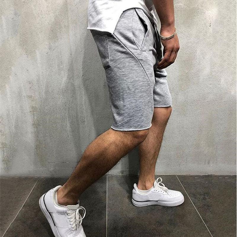 Man wearing casual grey shorts and white sneakers standing on dark tile floor against concrete wall