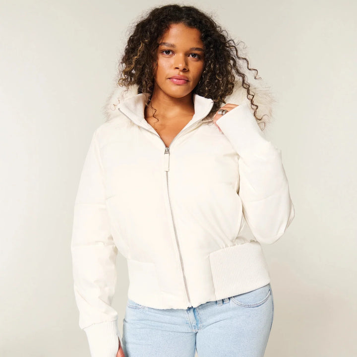 Woman wearing white puffer jacket with fur-trimmed hood and light blue jeans on beige background