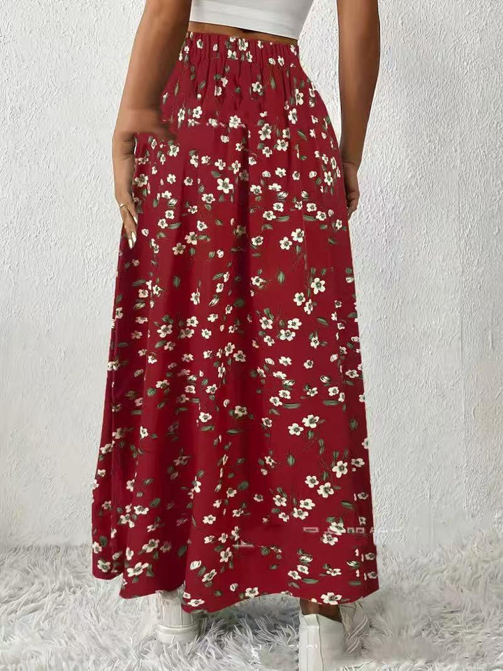 Back view of a woman wearing a red floral maxi skirt with white flowers and green leaves, paired with white shoes