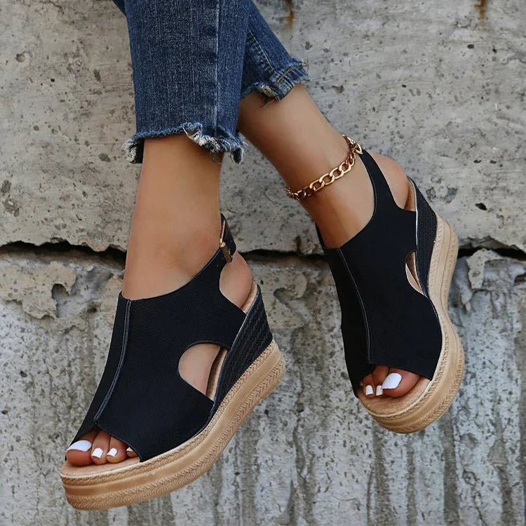 Black wedge sandals with cutouts and buckle worn with blue frayed hem jeans and gold anklet on stone background